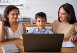 A boy is using his laptop at the kitchen table, flanked by two women. A notebook and a book lie nearby as they discuss how to join Google Classroom for parents, making sure everyone stays connected in their children's education.