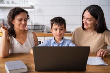 A boy is using his laptop at the kitchen table, flanked by two women. A notebook and a book lie nearby as they discuss how to join Google Classroom for parents, making sure everyone stays connected in their children's education.