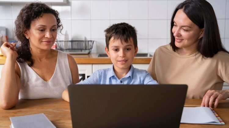 A boy is using his laptop at the kitchen table, flanked by two women. A notebook and a book lie nearby as they discuss how to join Google Classroom for parents, making sure everyone stays connected in their children's education.