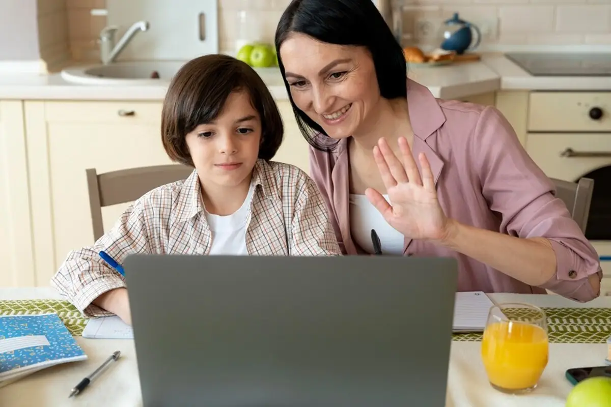 A woman and a child sit at a kitchen table, looking at a laptop screen. The woman is waving, as the Google Classroom App for Parents displays on the screen, while a glass of orange juice sits beside them.